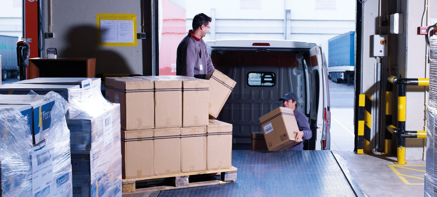 A imagem mostra duas pessoas a carregar ou descarregar caixas de uma carrinha num cais de carga.