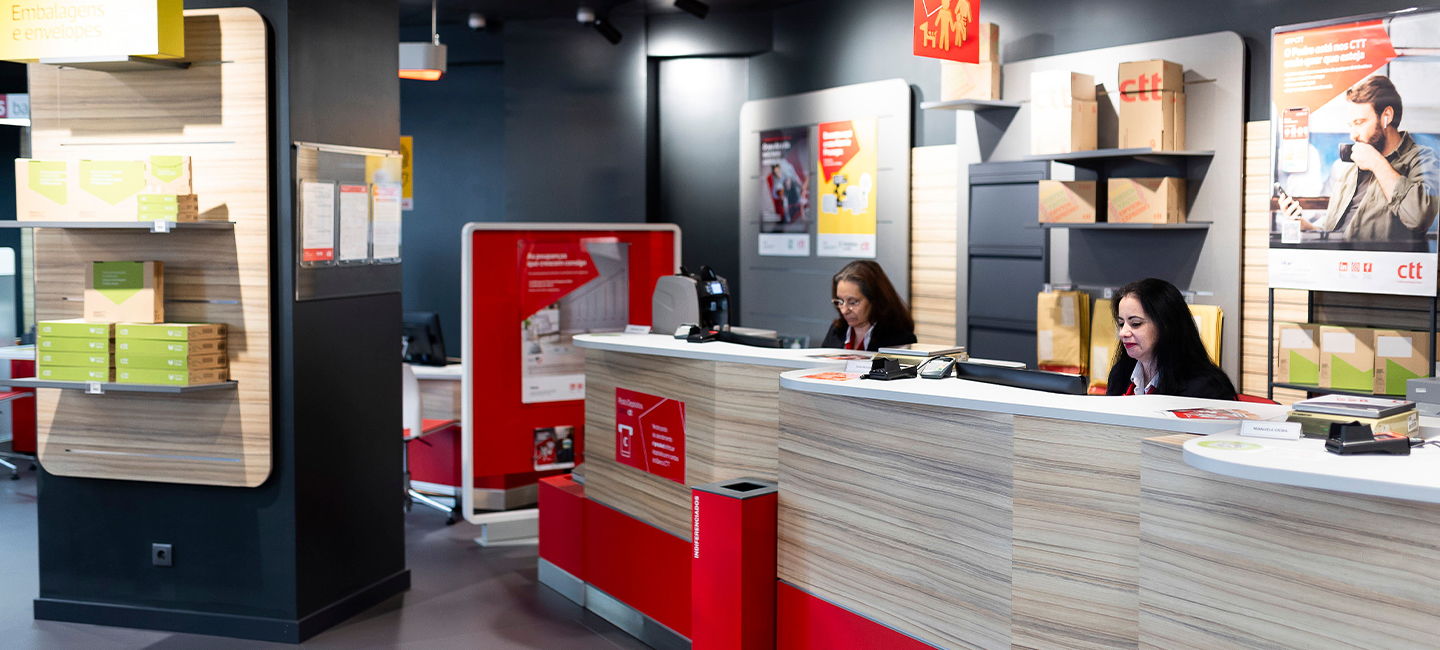 Senhoras que atendem o cliente nas lojas CTT, estão sentadas atrás do balcão de atendimento.