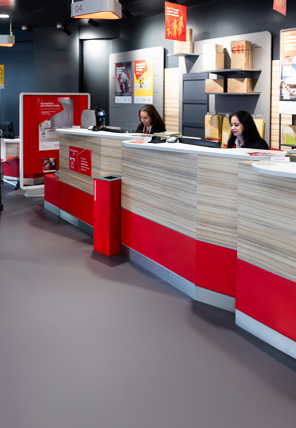 Senhoras que atendem o cliente nas lojas CTT, estão sentadas atrás do balcão de atendimento.